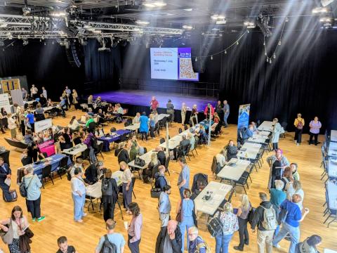 A bird's eye view of an event hall full of people, meeting at small desks and chatting. A stage and large screen can be seen in the background. 